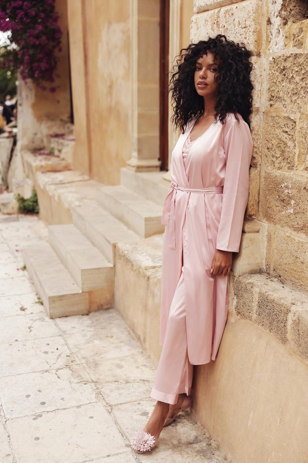 Eine Frau mit lockigem Haar trägt einen rosa Satinmantel und steht nachdenklich an einer Steinmauer